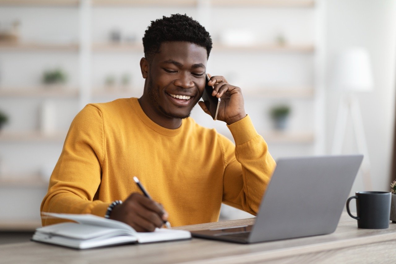 Man smiling while speaking on the phone and writing notes beside a laptop.